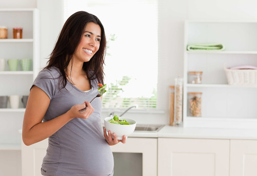 Beautiful pregnant woman holding a bowl of salad while standing in the kitchen