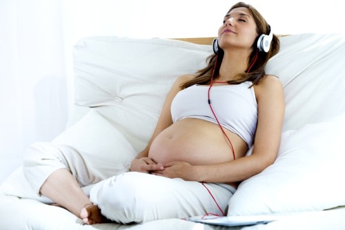 Portrait of pregnant woman sitting on sofa at home and listening music in headphones.