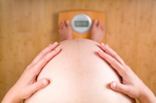 Pregnant woman weighing herself on a bathroom scale. View from above. No face. Focus on belly and hands.